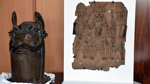 A tall, shiny bronze statue head with a patterned crown sits on the left. Next to it, on the right, is a flat bronze plaque showing three people standing together, wearing decorated clothes and holding objects