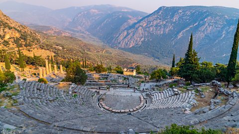 A big, ancient stone theatre sits on a hillside, shaped like a half-circle with many rows of seats. Behind it are tall, rocky mountains and lots of trees, all under a bright, hazy sky.
