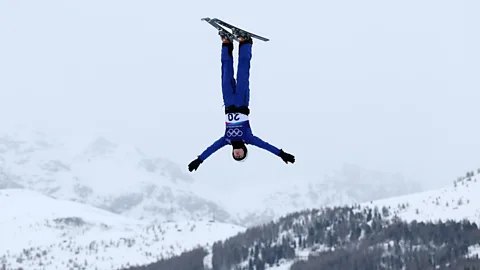 Getty Images A ski jumper mid-jump upside-down, with his arms outstretched and snowy mountains in the background