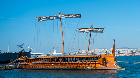 A large ship with two sails and ropes leading from them to the deck. At its sides there are many holes at three different layers where long oars poke out.