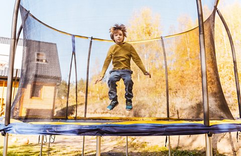 A boy wearing a yellow jumper and blue jeans jumping on a trampoline in a garden on a sunny day.
