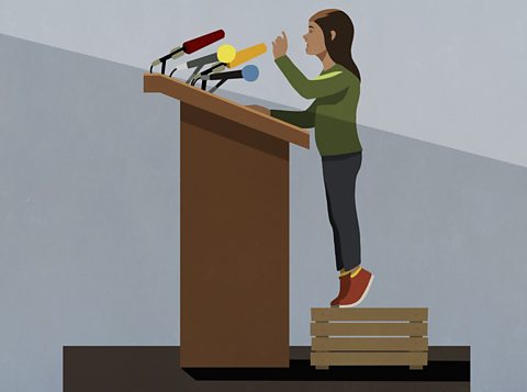 An image of a girl giving a speech, at a lecturn, with microphones, an example of persuasive language found on the SEAG transfer test