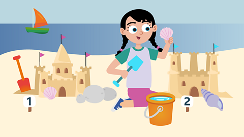 A girl on a beach holding a shell and a spade in between two sandcastles.