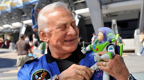 Astronaut Buzz Aldrin poses with Disney's Buzz Lightyear. He has white hair and wears a blue jacket and black top underneath. He smiles at the action figure he is holding, which has a green and white spacesuit, purple head cover and glass dome covering his head
