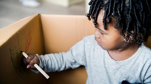 A young boy sits in a cardboard box, drawing with colouring pencils.