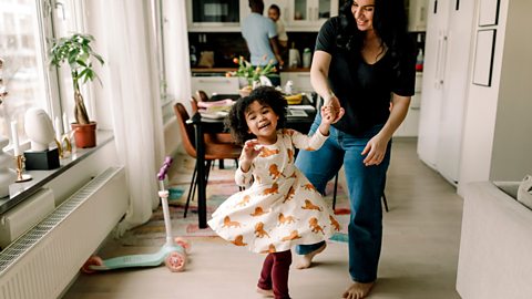 A young girl smiles, dancing with her mum in the living room.