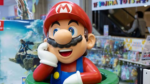 A large plastic figure of Nintendo character Mario sits in the middle of a busy toy story. Coming out of a green pipe, Mario - who has a moustache and wears a red cap, matching long-sleeved top and blue overalls - smiles and rests his arms on the pipe, with his head leaning on one palm