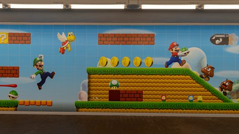 A poster in a subway showing an animated scene from a Mario Bros game. It shows characters Mario and Luigi, one in a red cap, long-sleeved top and blue overalls and the other matching in green, jumping in the air to collect gold coins and avoiding brown mushrooms with legs