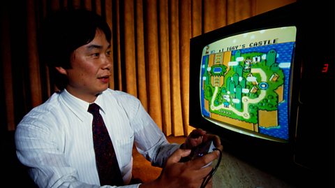 Creator Shigeru Miyamoto, who has dark thick hair, sits at a desk wearing a white shirt and black tie, playing Super Mario World on a Nintendo Super NES System. Holding a controller, on screen is a 90s-style game showing an island surrounded by sea and a mapped pathway
