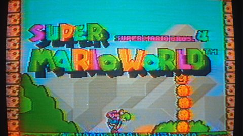 A pixelated screen shows Nintendo's Super Mario Bros. Mario World video game in 1991. It has bold, blocked letters in various colours and shows a dinosaur like green character carrying a backpack at the bottom of the screen