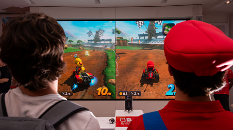 Customers, including a person dressed as Mario in a red cap on the left, play the Mario Kart World video game on a Nintendo Switch 2 console. On the duel screen is both player's characters driving their carts on a dirt track