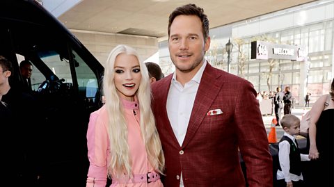 Anya Taylor-Joy and Chris Pratt attend a screening of The Super Mario Bros in 2023. Anya has long white hair and red lipstick, wearing a neck-high pink boiler suit. Chris has a burgundy blazer buttoned jacket with a white and red pocket square and a white shirt beneath