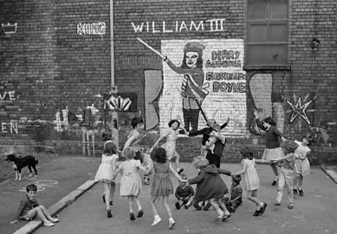 Children playing 'Ring a Ring o' Roses' on a street marked with sectarian graffiti, Belfast, 1962.