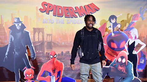 Shameik Moore at the Spider-Man: Into the Spider-Verse premiere. He stands in the centre wearing a black hoody and grey checked trousers. Behind him is a movie illustration poster depicting New York and around him are various cardboard cut-outs of different Spider-Man characters