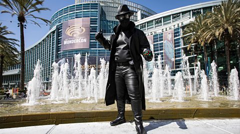 A Marvel fan dressed as Spider-Man Noir in front of a fountain outside of a hotel. Head to toe the costume is black, from a black hat and covering to leather pants and a long black trench coat