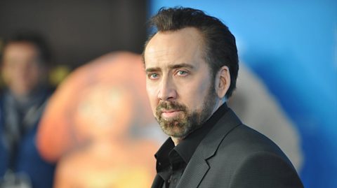 A close-up photo of actor Nicholas Cage at a film premiere. He looks at the camera and wears a black shirt and black suit jacket. He has brown hair and a brown beard