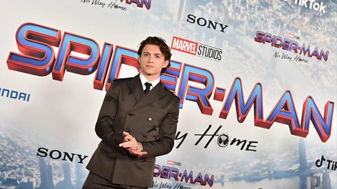 Actor Tom Holland at a Spider-Man premiere. He has longer brown curly hair and wears a grey suit with a white shirt and black tie
