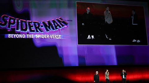 Spider-Man: Beyond the Spider-Verse appears on a screen in bold text with a purple background. On stage are three men discussing the upcoming film and talking into mics