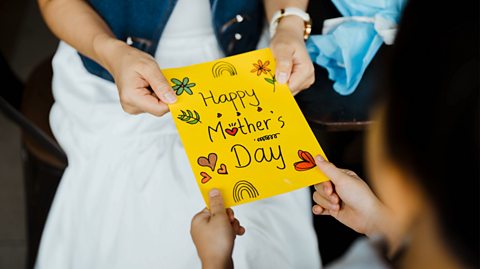 A close-up photo of a woman wearing a white dress and denim jacket being handed a handmade yellow card saying Happy Mother's Day with drawings of hearts, flowers and rainbows