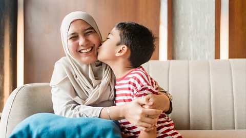 A woman wearing a cream headcovering sits on a cream couch hugging her young son. Her son kisses her cheek and wears and red and white striped top