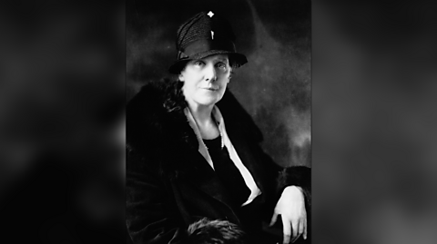 A black and white photo of Anna Jarvis who sits in a chair looking at the camera. She wears a black hat, a black coat with fur lining on the neck and cuffs and a white shirt and black top beneath