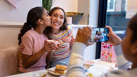 A little girl takes a photo on a camera phone of her mum and sister. They all sit at a table having afternoon tea for Mother's Day and there's a pink card on dis[lay on the table. The mum and daughter in the photo both hold pink mugs. The mum smiles and has a multi-coloured striped top and her daughter, who has dark hair in a ponytail kisses her cheek and wears a pink top