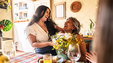 In a bright family kitchen, a adult daughter and her mum laugh and smile at one another. The daughter has long brown hair, a cream top and blue jeans and hands her mum a bouquet of bright flowers with yellow and white colours. The mum has a green top on and grey and black hair