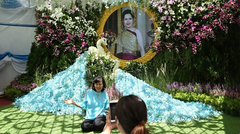 A woman poses in front of a portrait of Queen Sirikit during Queen Mother Sirikit's 88th birthday celebrations. The woman has a blue top, dark pants and black hair. Behind her is a shrine of blue flowers with a floral swan and the portrait with a gold frame. In the portrait, the Queen wears a gold and red dress