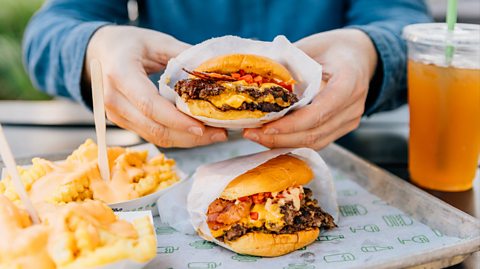 A man wearing a long-sleeved blue top holds a beef burger with cheese and bacon wrapped in white paper in their hands. Beneath is a cream tray with an identical burger on it and crimped fries with a light orange sauce over the top. Next to the tray is a cold orange drink in a plastic cup with a green straw