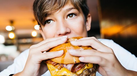 A close-up image of a boy with long brown hair and wearing a white top earing a beef with cheese and bacon