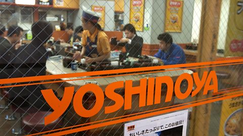 Yoshinoya, written in bold orange text, is printed on a window. Through the window, we can see diners inside the restaurant and a server wearing a white and navy cap, yellow top and dark pinafore
