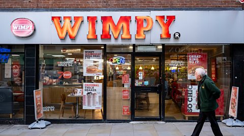 Outside a Wimpy burger site, with a grey shop front and text reading Wimpy in bold orange on a street. A man with grey hair and a green jacket walks past and looks at the shop window
