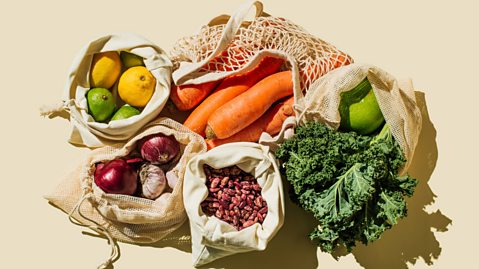A cream table with bags of fruit and vegetables on it, including lemons and limes, red onions, nuts, carrots and cabbage