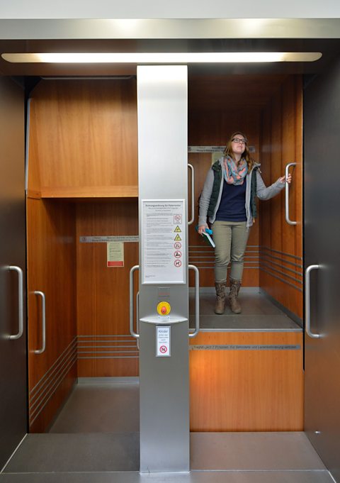 A woman holds onto a rail, stood inside the lift chamber, as a Paternoster lift moves in action