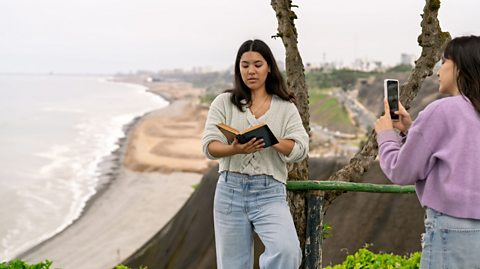 An image of a young woman reading outdoors while being filmed by a friend on her smartphone