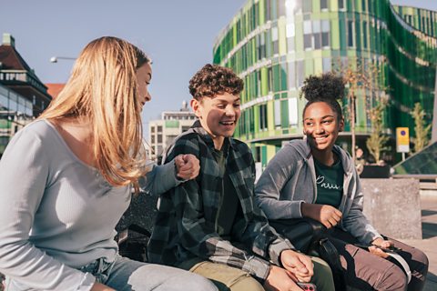 Group of student friends sitting together in a city outside in the sun with a green glass building in the background