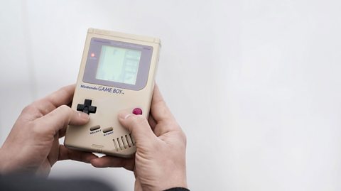 A shot of a Nintendo Game Boy, displaying a Tetris game in action, being held by two hands against a white backdrop