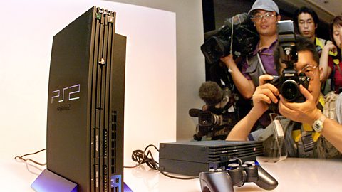 Two Sony Playstation 2 consoles and a controller being photographed by media in Japan at a console launch. One unit is stood vertically, the other horizontally