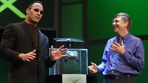 Dwayne 'The Rock' Johnson is stood next to the first Xbox and a controller on a plinth, with Bill Gates to the side laughing