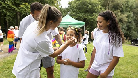 Two young girls wearing white t-shirts having rainbow face paints added by their mother.