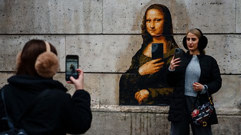 A woman taking a selfie, posing next to street art of the Mona Lisa depicted with a phone taking a selfie, while another woman takes a picture of them