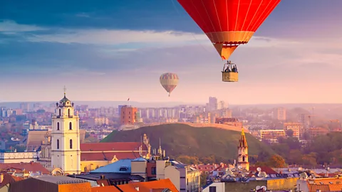 Getty Images Hot air balloons floating above Vilnius Old Town with church tower, red rooftops and Gediminas Hill visible below (Credit: Getty Images)