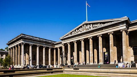 An image of the British Museum. A big, old stone building with tall columns in front. People are walking and sitting outside on a sunny day, and a Union Jack waves on top of the roof.