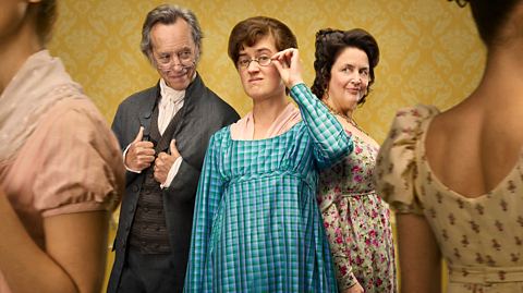 Richard E Grant, Ella Bruccoleri and Ruth Jones in BBC's The Other Bennet Sister. Grant has a grey suit on, grey hair and small glasses. Bruccoleri has brown hair tied up, small circular glasses and a long sleeved blue dress on. Jones has curly dark hair styled up with a floral long sleeved dress on. They all stand against a yellow background