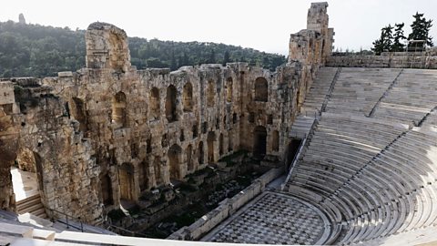 A big, old stone theatre with rows of curved seats outdoors. The stage area is made of ancient rocks and walls.