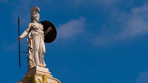 A tall stone statue of Athena holding a long spear and a round shield standing on top of a decorated pillar. 