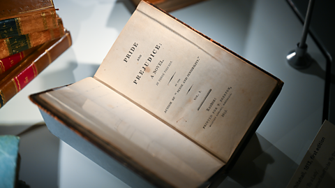 A first edition of the book Pride and Prejudice is displayed at Jane Austen's House on a white table, with a pile of leather bound books next to it. It is open to the old front page which reads 'Pride and Prejudice - a novel'