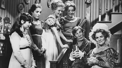 A black and white photo of actors playing the Bennet family. Four daughters in long dresses with frills stand on a staircase. In the hall is their mother smiling wearing a puffy bonnet and a ruffled dressed and their father holding a book looking at her