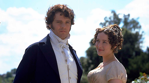 Colin Firth and Jennifer Ehle in character as Mr. Darcy and Elizabeth Bennet. On a sunny day, they stand in gardens, Firth looking at the camera and Ehle smiling looking at Firth. First has a navy blue coat and cream waistcoat, while Ehle wears a short sleeved champagne coloured dress and has her curly brown hair styled up