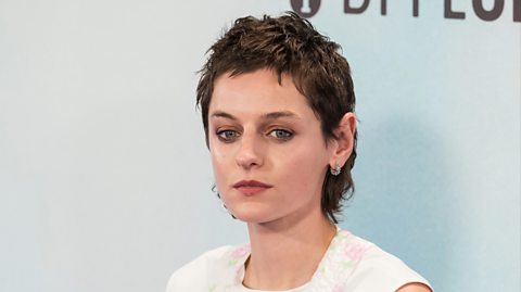 Actress Emma Corrin at a film premiere. The close-up photo shows Corrin looking at the camera. She has short brown hair and wears a white and floral top